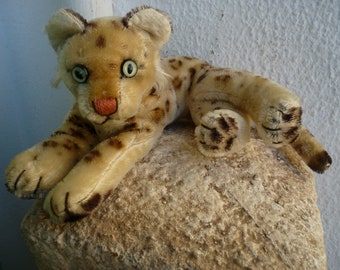 Könnte beinhalten: Ein Plüsch-Leopard-Spielzeug mit braunem und braunem Fell, großen grünen Augen und einer rosa Nase. Das Spielzeug liegt auf einer rauen, strukturierten Oberfläche.