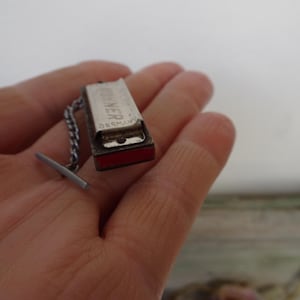 May include: A miniature silver-toned harmonica tie tack with a chain and bar closure. The harmonica has the word "HOHNER" printed on it, with "GERMANY" below. The bottom of the harmonica is red.