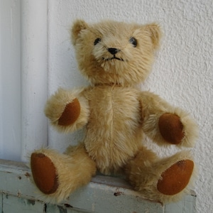 Op de afbeelding: Een vintage teddybeer met bruine vacht en bruine viltpootjes. De beer zit op een houten oppervlak.