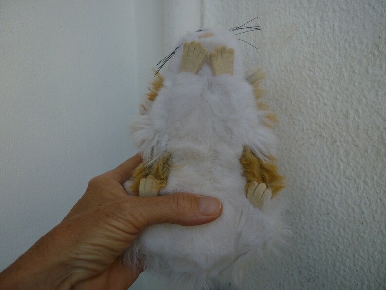 May include: A white and brown plush toy hamster with black eyes and whiskers. The hamster is lying on its back with its paws in the air.