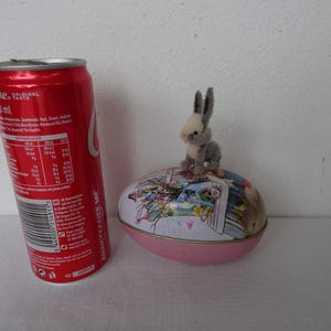 Puede incluir: Una lata roja de Coca-Cola junto a una caja decorativa de huevos de Pascua con una pequeña figura de conejito gris encima. El huevo es rosa con un diseño floral y un borde dorado.