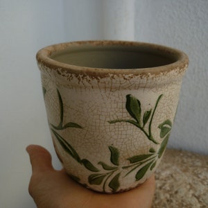 以下が含まれることがあります： 緑色の花柄が描かれた、白の陶器製のプランター。プランターは、開口部が広く、丸みを帯びており、縁が少し広がっています。