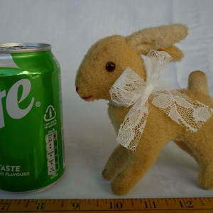 May include: A tan plush toy deer with a white lace bow. The deer has brown eyes and a pink nose. The toy is standing on a wooden ruler.
