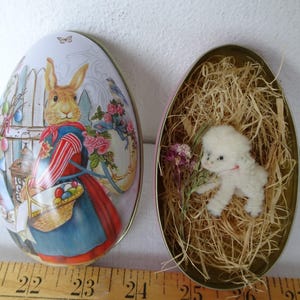 Puede incluir: Un recipiente de hojalata en forma de huevo con una escena de Pascua pintada que presenta un conejo sosteniendo una canasta de huevos. La otra mitad del recipiente contiene una pequeña figura de cordero blanco anidada en paja. Un artículo decorativo con temática de Pascua.
