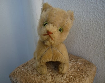 Puede incluir: Un gato de peluche vintage con pelaje marrón, ojos verdes y una nariz cosida. El gato está sentado sobre una superficie texturizada.