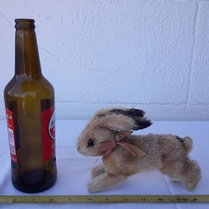 May include: A brown and tan stuffed rabbit toy with a brown bow around its neck. The rabbit is sitting on a white surface next to a brown glass bottle.