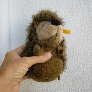 May include: A brown and black plush hedgehog toy with a yellow plastic nose. The hedgehog is being held by a hand.