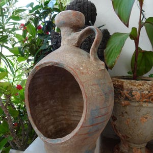 Op de afbeelding: Een rustieke terracotta pot met een grote ronde opening aan de voorzijde, een smalle hals en een enkel handvat aan de zijkant. Het oppervlak heeft een verweerde, aardse textuur met vage sporen van blauwe en rode pigmentaccenten.