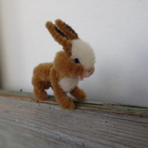 Puede incluir: Un pequeño conejo marrón y blanco hecho de hilo. El conejo tiene una cara blanca y pelaje marrón en su cuerpo. Está de pie sobre una superficie de madera.
