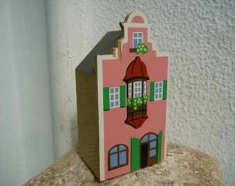 May include: A pink wooden house with a green roof and white trim. The house has a balcony with a red railing and green plants. The windows are blue and the door is green.