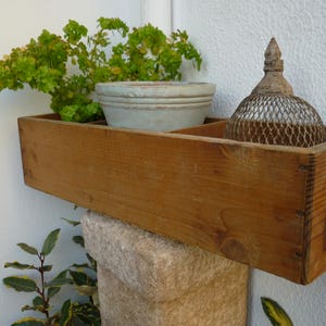 Può includere: Un vaso da fiori in legno con una pianta verde chiaro e un vaso in ceramica bianca. Il vaso da fiori è appoggiato su una superficie in pietra. Una gabbia di metallo decorativa è anche nel vaso da fiori.