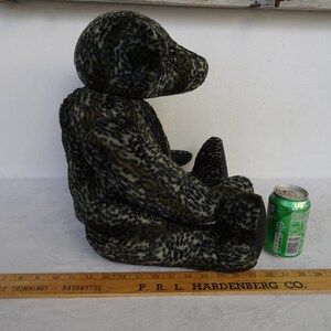 May include: A plush teddy bear in a seated position, featuring a mottled pattern of black and olive green. The bear has black paws and a small, round head. A green soda can is next to the bear for size comparison.