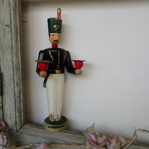 Puede incluir: Una figurita de madera de un soldado que lleva un uniforme negro y verde con un sombrero rojo y blanco. El soldado sostiene dos candelabros en sus manos. La figurita es una decoración navideña alemana tradicional.