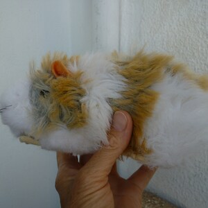 May include: A plush toy hamster with white, brown, and gray fur. The hamster has a small orange ear and is being held by a hand.
