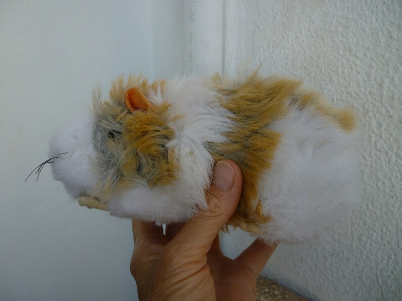May include: A plush toy hamster with white, brown, and gray fur. The hamster has a small orange ear and is being held by a hand.