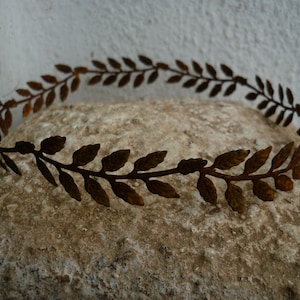 May include: A bronze-colored metal leaf crown, resting on a textured stone surface. The crown features a repeating pattern of stylized leaves, creating a circular design. The metal has a rustic, aged appearance.