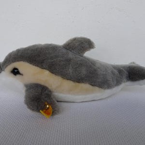 Può includere: Un peluche a forma di delfino nei toni del grigio, beige e bianco. L'animale di peluche ha una piccola pinna, un'etichetta gialla e occhi neri. Il delfino è su una superficie bianca.