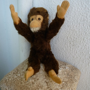 May include: A brown plush monkey toy with a tan face and large, expressive eyes. The monkey is sitting with its arms raised in the air, as if it is waving.
