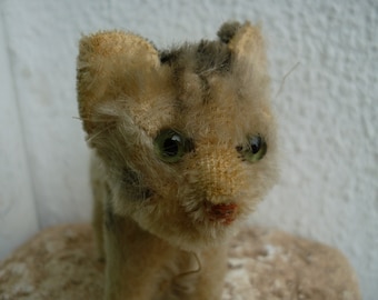 May include: A vintage plush toy cat with brown and gray fur, green eyes, and a pink nose. The cat is sitting on a light brown surface.