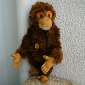 May include: A brown plush monkey toy with a light brown face and a red ribbon around its neck. The monkey is wearing a gold tag with the word "Steiff" on it.