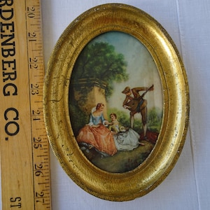 Pode incluir: Pintura oval antiga emoldurada em ouro. A obra retrata uma cena pastoral com três figuras em trajes de época, contra um fundo de árvores e uma estrutura de pedra. A moldura apresenta sinais de idade.