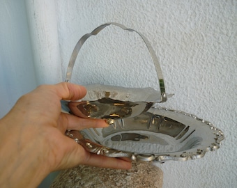 May include: A silver-colored serving basket with a handle, featuring a scalloped edge and ornate detailing. The basket is held up, showcasing its reflective surface and design. The basket is likely used for serving food or displaying items.