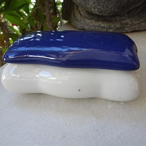 Può includere: Un portagioie in ceramica con finitura lucida. Il coperchio è blu scuro e la base è bianca. La scatola ha una forma arrotondata e organica, che suggerisce una sensazione liscia e tattile. La scatola è appoggiata su una superficie bianca.