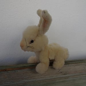 Może przedstawiać: Pluszowa zabawka w kształcie kremowego królika z prostymi uszami i czarnymi wąsami. Królik jest umieszczony na drewnianej powierzchni na tle białej ściany. Zabawka ma miękki, teksturowany wygląd.