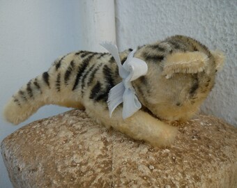 May include: A plush toy cat with black and white stripes, wearing a light blue bow. The cat is lying on a rough, brown surface.