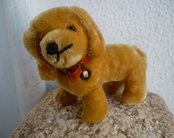 Puede incluir: Un perro de peluche marrón con un collar rojo y una campana. El perro está sentado sobre una superficie texturizada.