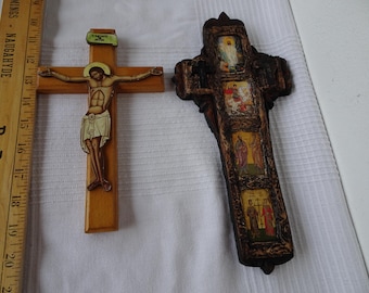 May include: Two wooden crosses. One is a light brown crucifix with a painted figure of Jesus. The other is a dark brown, ornate cross with several painted religious scenes. A wooden ruler is on the left side of the image.
