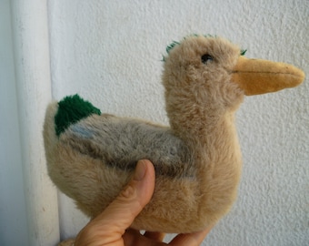 Puede incluir: Un pato de peluche con un cuerpo marrón y beige, una cabeza verde y un pico amarillo. El pato está sentado en una mano.