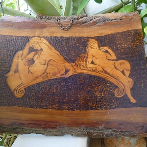 Peut inclure: Une plaque en bois avec un motif de deux figures allongées sur une surface courbe, réalisé à l'aide de bois brûlé. La plaque est suspendue à une chaîne.