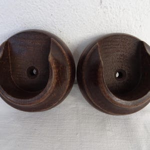 May include: Two circular dark brown wooden curtain rod brackets. Each bracket features a deep, U-shaped cutout at the center designed to hold a rod, with a small, centered circular hole for mounting hardware. The wood grain is visible throughout.