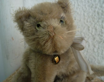 Puede incluir: Un juguete de gato de peluche cubierto de pelaje castaño claro y desgreñado con ojos de plástico verde, bigotes largos y una cinta de color crema atada alrededor del cuello. Un pequeño dije de metal dorado con un centro oscuro está unido a la cinta.