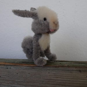 May include: A small, plush bunny rabbit toy. The rabbit is primarily gray with a white chest and head. It has black eyes and a pink nose. The toy is sitting on a wooden surface.