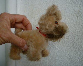 Puede incluir: Un pequeño perro de peluche marrón con una cinta roja alrededor del cuello. El perro tiene pelo marrón y una nariz negra.