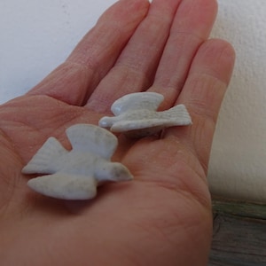 Puede incluir: Dos pequeñas figuras de pájaros de cerámica en color blanco roto con las alas extendidas. Cada pájaro presenta un acabado mate y liso con una forma sencilla y estilizada. Las figuras están representadas en posición de vuelo con detalles tallados.