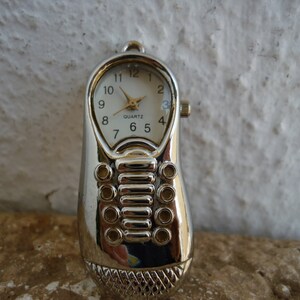 Könnte beinhalten: Silberfarbene Taschenuhr in Form eines Mobiltelefons mit weißem Zifferblatt und schwarzen Zeigern. Das Zifferblatt der Uhr hat schwarze Zahlen und die Aufschrift "QUARTZ" in goldenen Buchstaben.