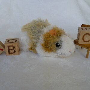 May include: A plush toy hamster with brown and white fur, lying on a white surface. Three wooden blocks with the letters A, B, and C are in front of the hamster. A small wooden chair with the letter C on the back is to the right of the hamster.