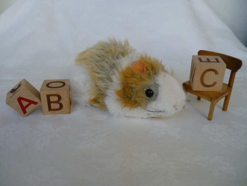 May include: A plush toy hamster with brown and white fur, lying on a white surface. Three wooden blocks with the letters A, B, and C are in front of the hamster. A small wooden chair with the letter C on the back is to the right of the hamster.