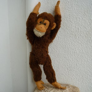 May include: A brown plush monkey toy with a light brown face and large, expressive eyes. The monkey is standing on its hind legs with its arms raised above its head.
