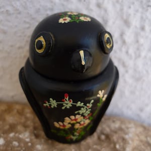 Può includere: Figurina di gufo in ceramica nera con fiori dipinti in bianco e giallo e bacche rosse. Il gufo ha un becco nero e occhi gialli.