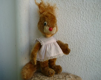 Puede incluir: Un juguete de peluche marrón en forma de ardilla que lleva un vestido rosa con ribete de encaje blanco. El juguete tiene la nariz roja y ojos grandes y negros.