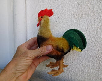May include: A plush toy rooster with a red comb and wattle, brown and yellow fur, and a green tail. The rooster is standing on a white surface.