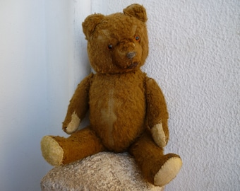 Puede incluir: Un oso de peluche marrón muy querido con ojos de botón se sienta sobre una superficie de piedra. El oso tiene una textura peluda desgastada y le falta algo de pelaje.