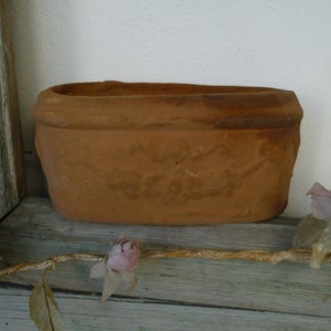 May include: An oval-shaped terracotta planter with a rustic, unglazed finish and a thick, rounded rim. The exterior surface features subtle, embossed decorative patterns that wrap around the sides of the vessel, showcasing a natural, earthy brown tone.