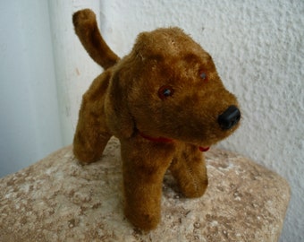 May include: A vintage brown plush dog toy with a red collar and black nose. The toy dog has small, round, blue eyes and is standing on a stone surface. The dog's fur is a warm, medium brown color.