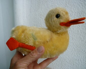 Puede incluir: Un juguete de peluche de pato amarillo con un pico y patas naranjas. El pato está sentado en una mano.