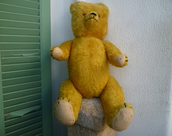 Puede incluir: Un oso de peluche amarillo vintage con un diseño clásico. El oso tiene una textura suave y afelpada, con patas color canela y una nariz y boca cosidas en negro. Está sentado sobre un pedestal de piedra.